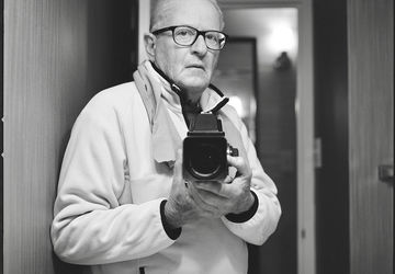 A man in glasses holding a camera chest-level, photographing himself in a mirror.
