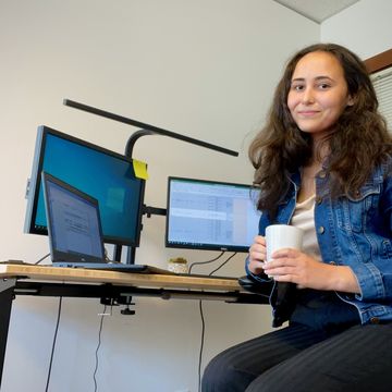 Tida Rau sits at her desk holding a mug while looking at the camera smiling.