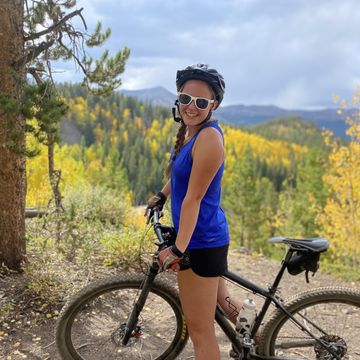 A student on a mountain bike with a helmet and sunglasses smiles at the camera with the mountains of Colorado in the background.