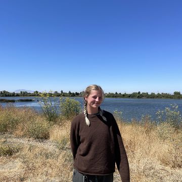 A young woman with blond braids stands in front of a body of water wearing a brown sweater and sweat pants squinting into the camera and smiling.