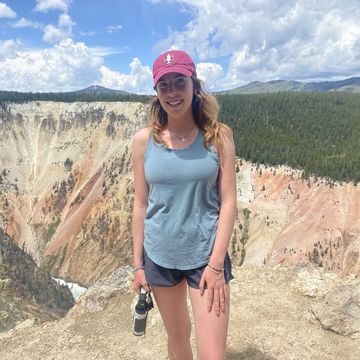 Elisabeth Westerman stands in a tank top, shorts and a red cap with the Grand Canyon and blue sky in the background.