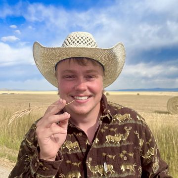 A young man in a brimmed hat and a brown button-down shirt smiles at the camera with a piece of wheat between his teeth and blue sky behind him