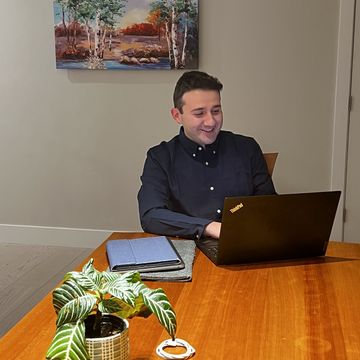 A young man in a button-down blue shirt works at a table on his laptop.