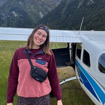 Full-body photo of Serena Turner standing in front of an airplane with mountains in the background, wearing a pink and purple fleece zip-up hoodie and dark patterned leggings.