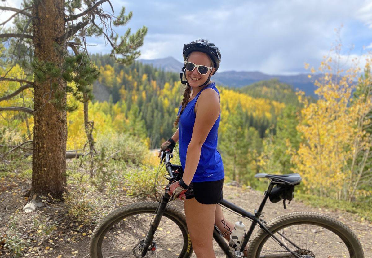 A student on a mountain bike with a helmet and sunglasses smiles at the camera with the mountains of Colorado in the background.