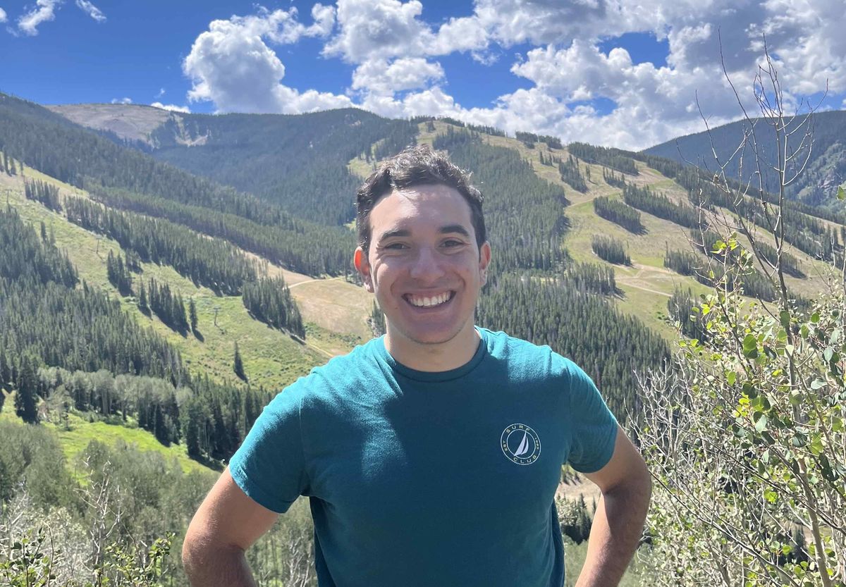 Lee Rosenthal in a green t-shirt smiles at the camera against a backdrop of blue sky and green rolling hills.