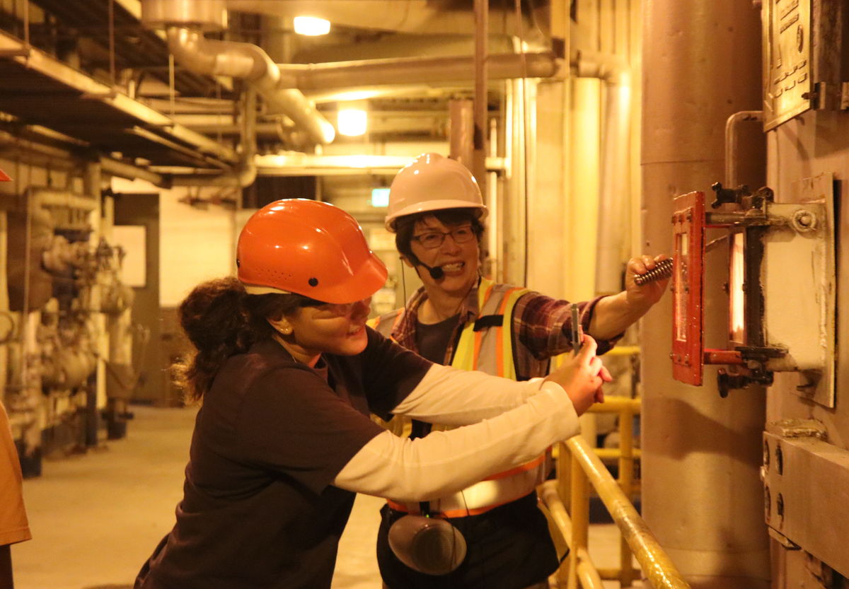 A girl in an orange hard hat leans forward and inspects something at the Spokane Waste to Energy Facility as an employee looks on