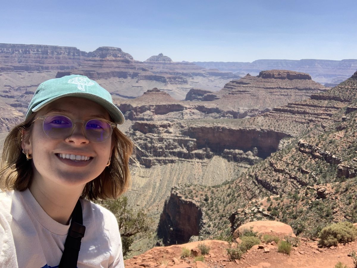 Against a backdrop of the South Rim of the Grand Canyon, Carly Taylor smiles at the camera wearing a blue baseball cap